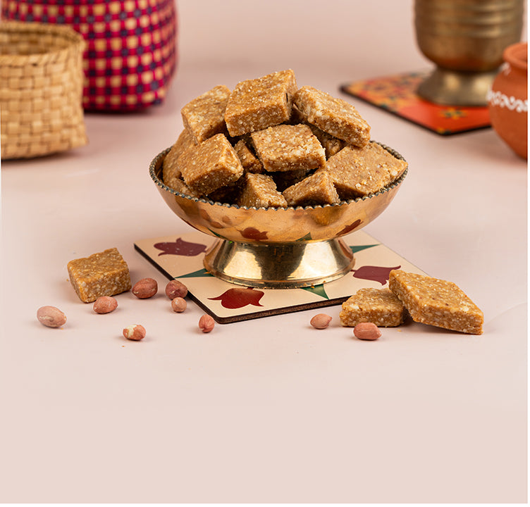 A bowl of Kovilpatti Kadalai Barfi, also known as peanut barfi, on a table with some scattered peanuts around it. The background includes a woven mat and various decorative items.