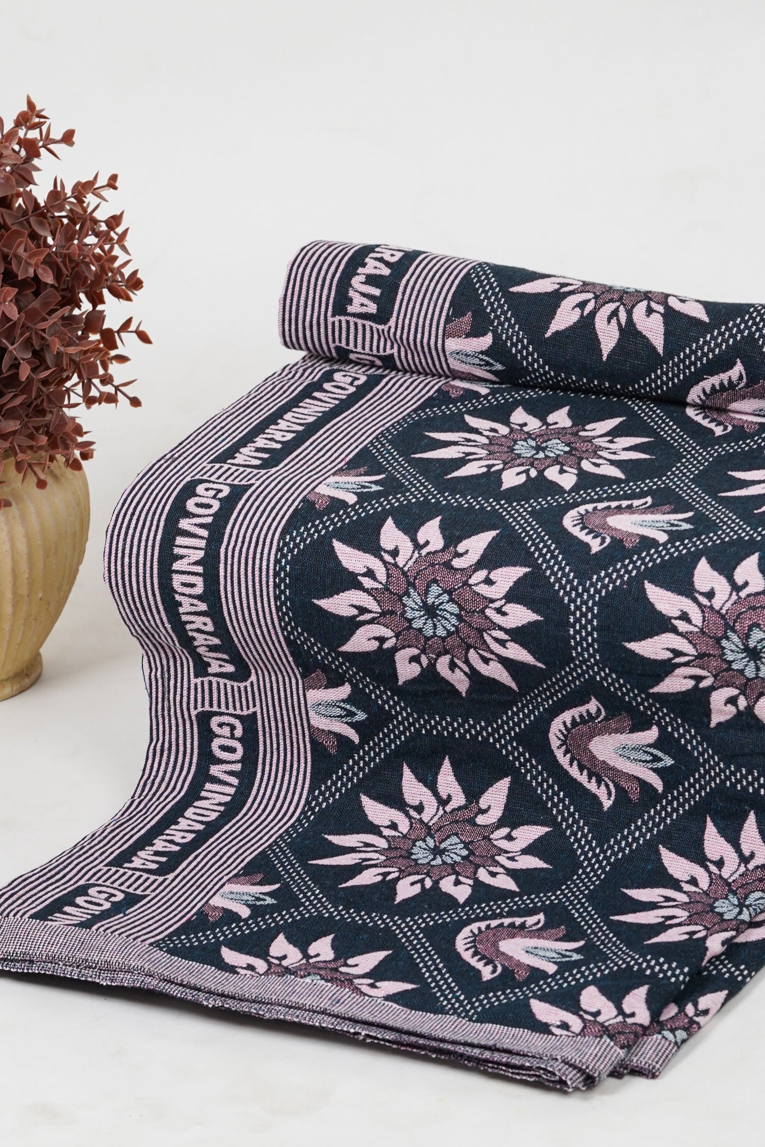 Dark blue fabric with pink floral patterns and striped borders, partially rolled and displayed on a light surface beside a small potted plant with reddish leaves.
