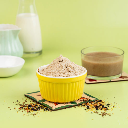 A yellow bowl filled with a beige health mix powder, with scattered ingredients around it. In the background, there's a glass bottle and a cup with a brown liquid, possibly the mixed beverage.