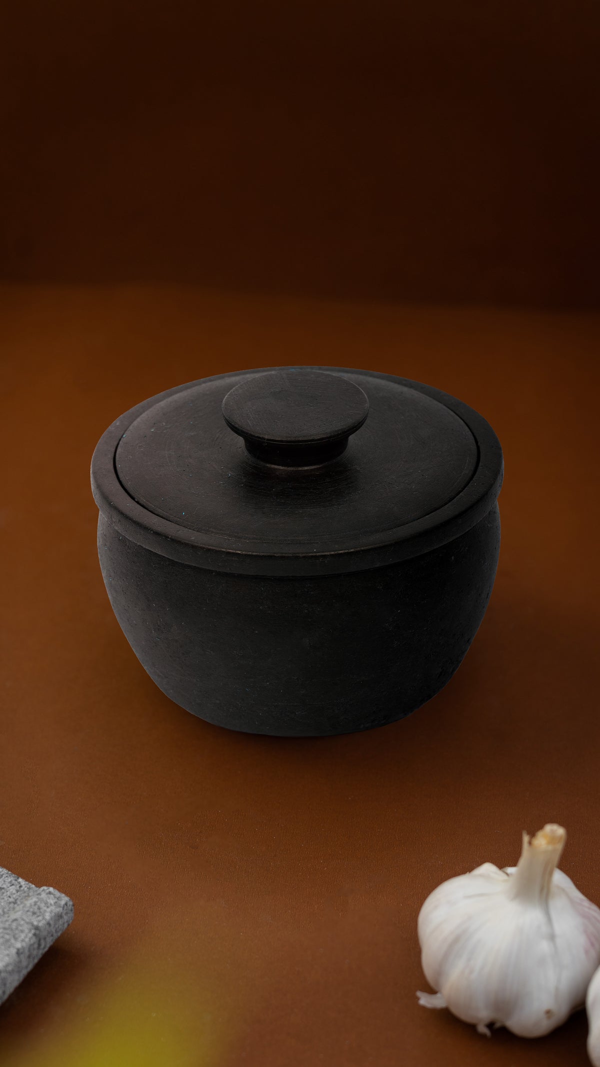 Black ceramic pot with a lid on a brown surface with garlic and a stone.
