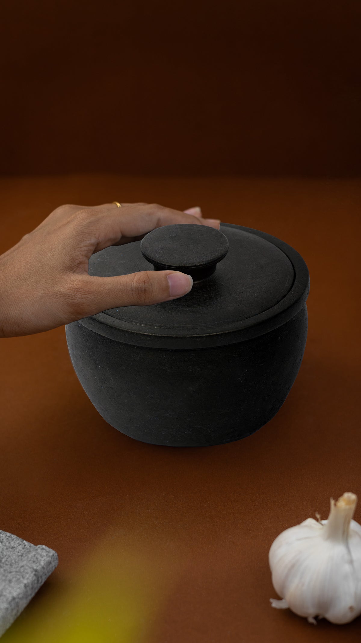 Person holding a black ceramic container on a brown surface with garlic and a stone.