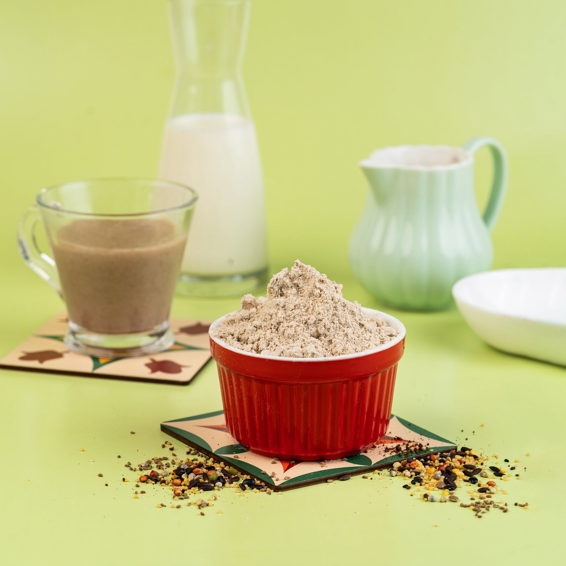 A bowl of 48-Ingredient Health Mix placed on a table with a glass of milk and a teapot in the background, alongside various scattered seeds and grains.