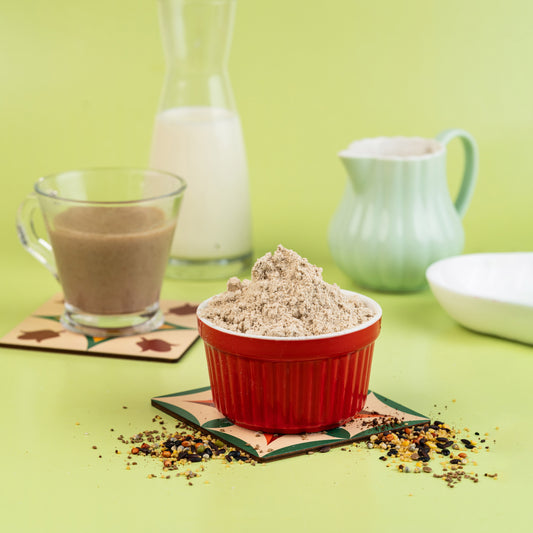 A bowl of 48-Ingredient Health Mix placed on a table with a glass of milk and a teapot in the background, alongside various scattered seeds and grains.