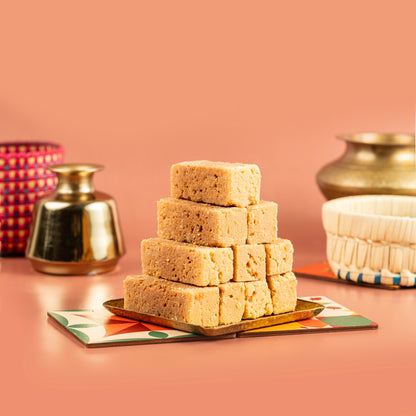 A stack of golden-brown Mysore pak sweets on a small round plate with a beige base, placed against a pink background with decorative items in the background.