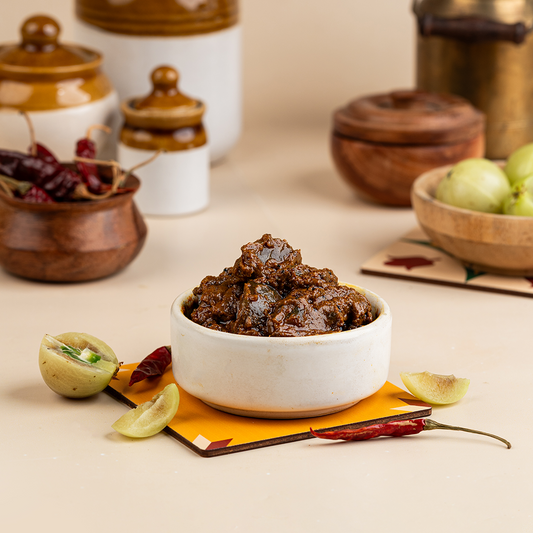 A white bowl filled with amla (Indian gooseberry) pickle, placed on a yellow napkin. The background includes a variety of kitchen items and some whole amla fruits.