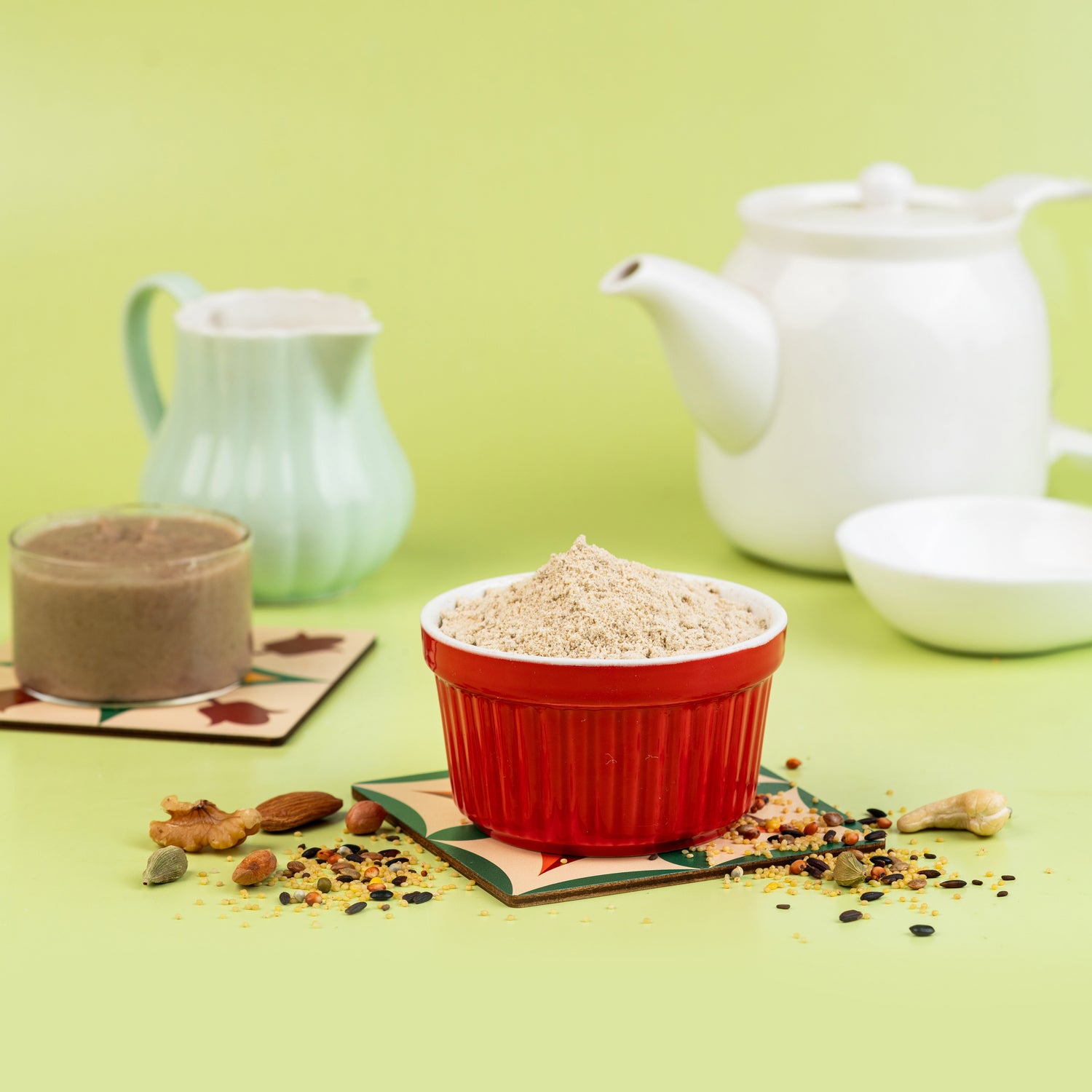 A red bowl of baby food with multigrain, placed on a green mat, with a white teapot and a blue cup in the background.