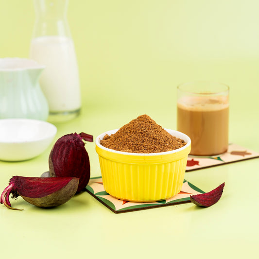 A bowl of beetroot malt powder is displayed in front of a glass of prepared beetroot malt drink and a raw beetroot on a yellow background.