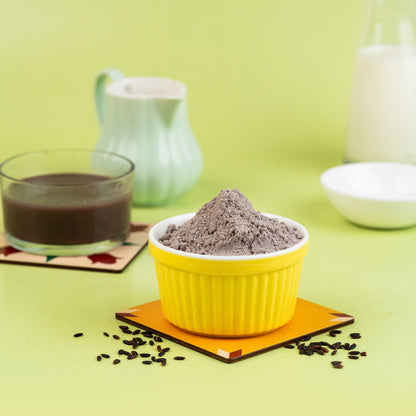 A yellow bowl filled with black rice porridge, a glass of milk, and a bowl of white porridge in the background, all placed on a green surface with coffee grounds scattered around.