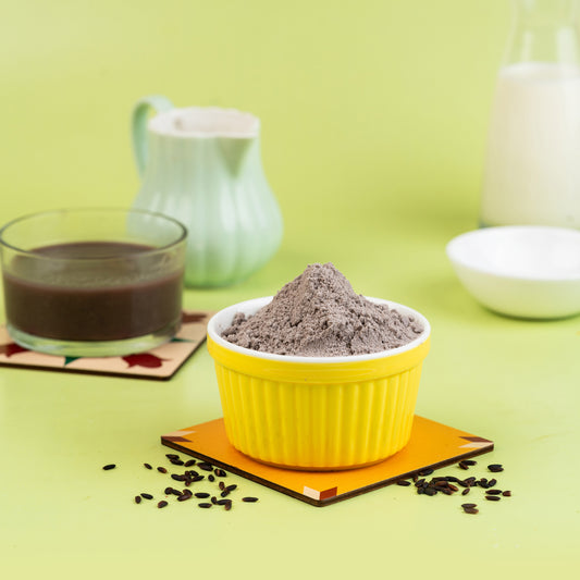 A yellow bowl filled with black rice porridge, a glass of milk, and a bowl of white porridge in the background, all placed on a green surface with coffee grounds scattered around.