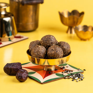 A bowl of black urad dal and dates ladoos on a yellow surface, with spices and a small bowl in the background.