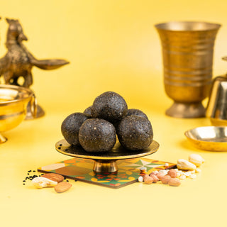 A plate with three round calcium Ladoos, placed on a yellow surface with a backdrop of metal bowls and a decorative pattern. The Ladoos are black in color.