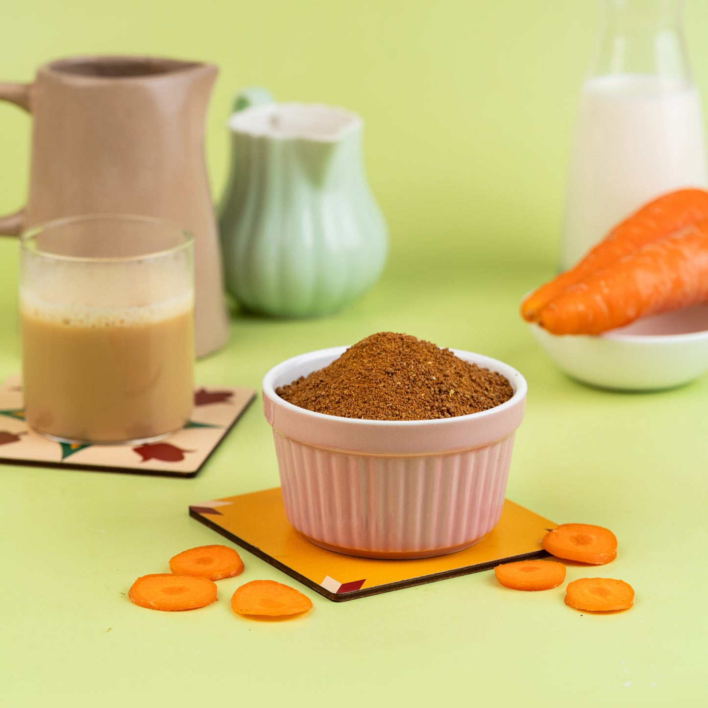 A bowl of carrot malt powder is displayed in front of a prepared carrot and a glass of milk, on a green background with decorative coasters and a pitcher.