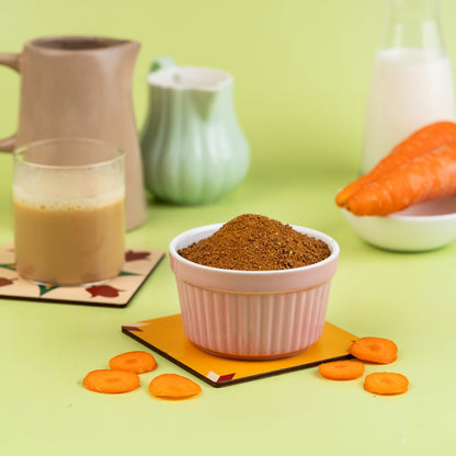 A bowl of carrot malt powder is displayed in front of a prepared carrot and a glass of milk, on a green background with decorative coasters and a pitcher.