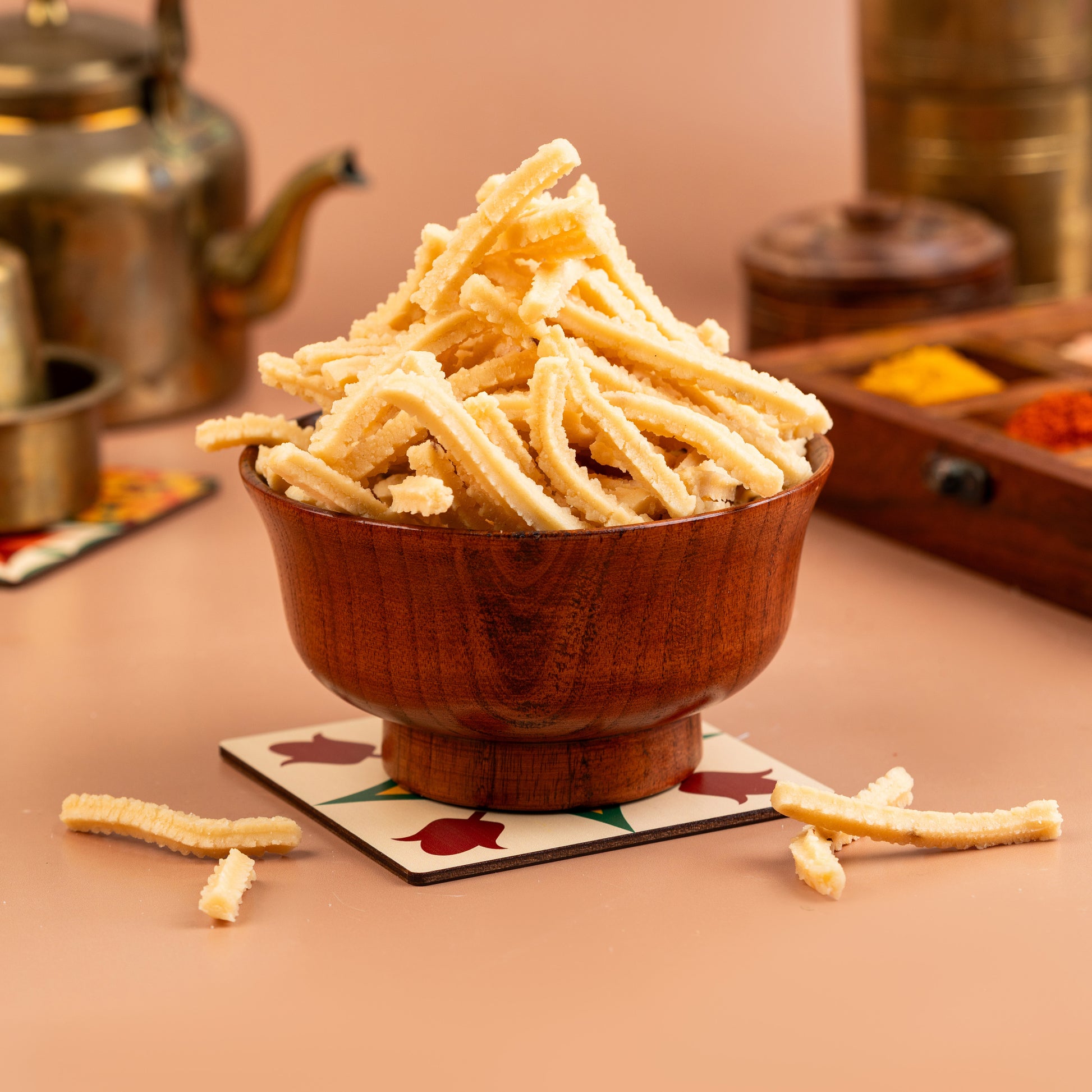 A wooden bowl filled with Chettinad Butter Murukku, a crispy snack, placed on a placemat with a tea set in the background.