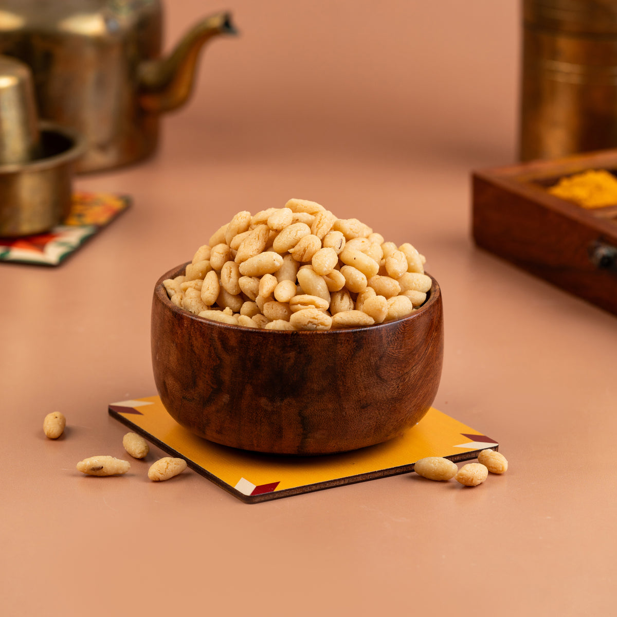 A wooden bowl filled with small, crispy golden-brown trail mix balls, placed on a copper-colored plate with a backdrop of various spices and a teapot in the background.