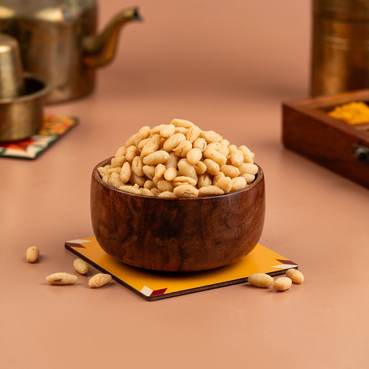 A wooden bowl filled with small, crispy golden-brown trail mix balls, placed on a copper-colored plate with a backdrop of various spices and a teapot in the background.
