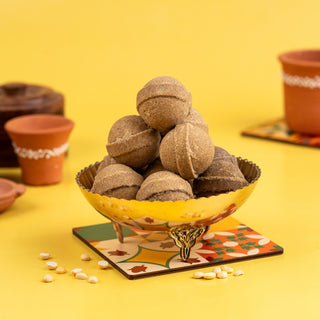 A bowl of Chickpeas Ghee Ladoo on a yellow plate with a beige border, with potted plants and millet grains scattered around.