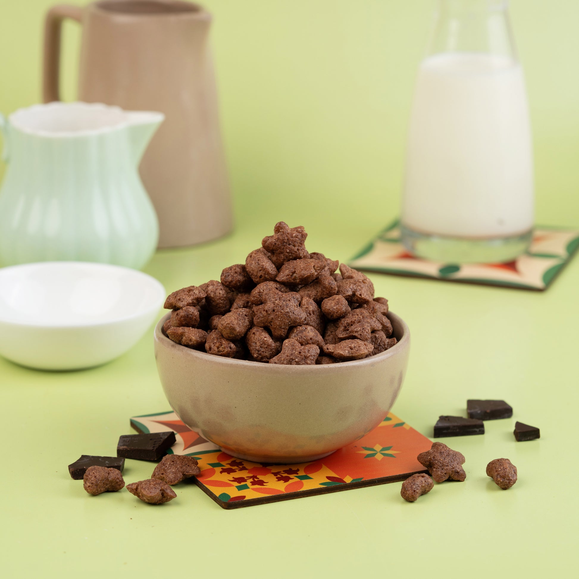 A bowl of Choco Millets on a colorful placemat, with a jug of milk and a bowl in the background, all placed on a green surface.