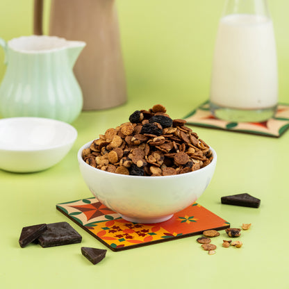 A bowl of Choco Muesli which includes crunchy oats, chocolate chunks, and nuts, placed on a colorful coaster with milk and chocolate pieces around it.