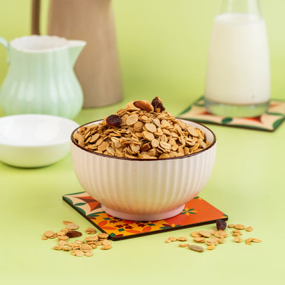 A bowl of classic muesli made of crunchy oats, nuts, and dried fruits, placed on a colorful coaster with a bottle of milk and a cream-colored bowl in the background.