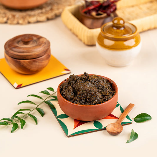 A bowl of Curry Leaf Thokku (Karuvepillai) served on a colorful placemat, with a wooden spoon and a small jar in the background, all set against a traditional Indian backdrop.