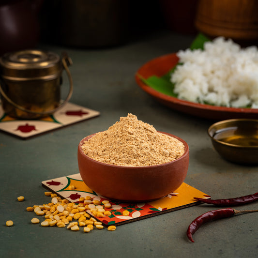 A bowl of Paruppu Podi (spice mix) on a colorful fabric, with a background of rice and a red spice container, set on a table with a South Indian patterned placemat.