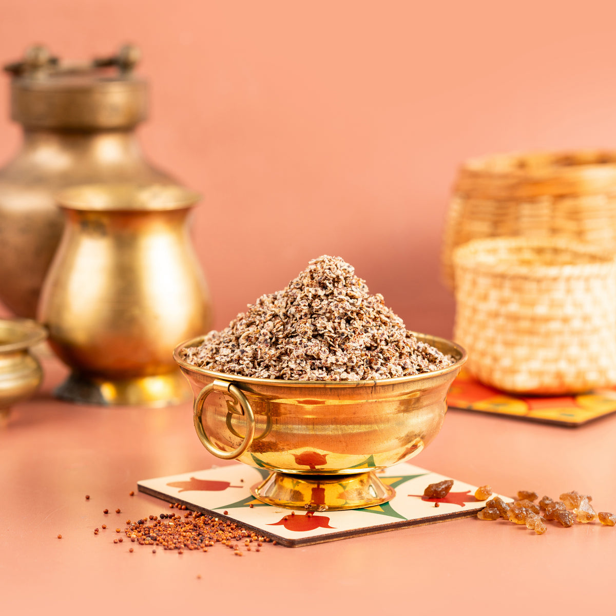 A bowl of air fried ragi sweet on a plate with a backdrop of woven baskets and a golden pot. The ragi is sprinkled with spices and sits in a square, decorative plate with a pink background.