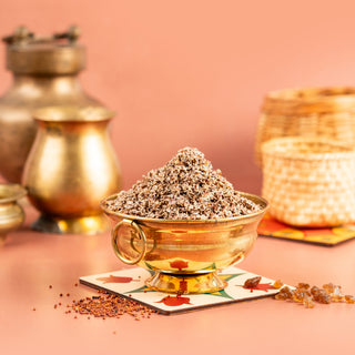 A bowl of air fried ragi sweet on a plate with a backdrop of woven baskets and a golden pot. The ragi is sprinkled with spices and sits in a square, decorative plate with a pink background.