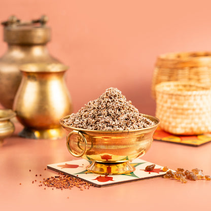 A bowl of air fried ragi sweet on a plate with a backdrop of woven baskets and a golden pot. The ragi is sprinkled with spices and sits in a square, decorative plate with a pink background.