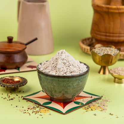 A green bowl filled with Five Millet Porridge Mix, with a spoon resting in the bowl. The background includes a beige pot, a wooden spoon, and a patterned tablecloth.