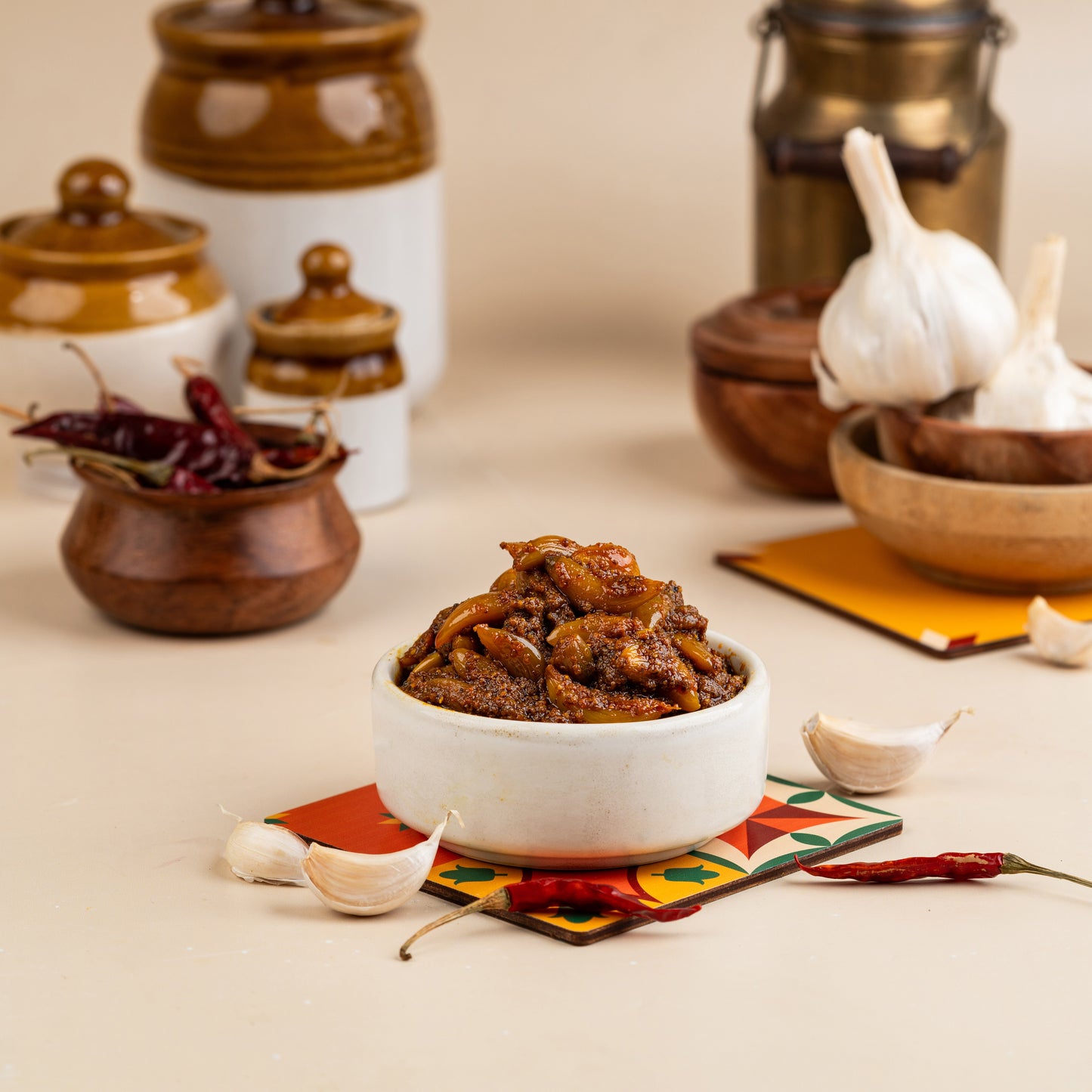 A white bowl filled with garlic pickle, placed on a colorful placemat. In the background, there are two jars and a piece of garlic on a spoon.