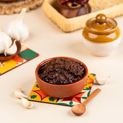 A bowl of Garlic Thokku (Poondu) on a colorful mat, with garlic cloves and a wooden spoon beside it.