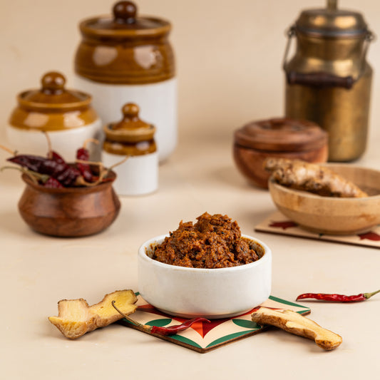 A bowl of ginger pickle in the foreground with clay pots in the background, along with some whole spices like cinnamon sticks and cardamom pods.