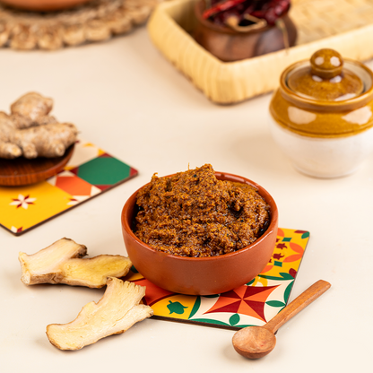 A bowl of ginger thokku (inji) with fresh ginger roots and a spoon on a colorful placemat, against a backdrop of various spices and a pottery object.