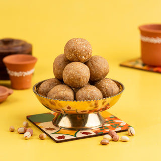 A golden bowl filled with groundnut ghee ladoos, placed on a colorful plate with a backdrop of yellow and beige pottery. Some peanuts are scattered around the bowl.