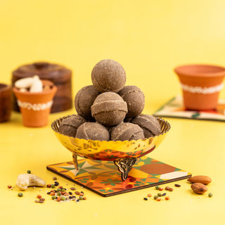 A brass bowl filled with brown millet ladoos (Sathu Maavu) on a colorful mat, with nuts and decorative elements around it.