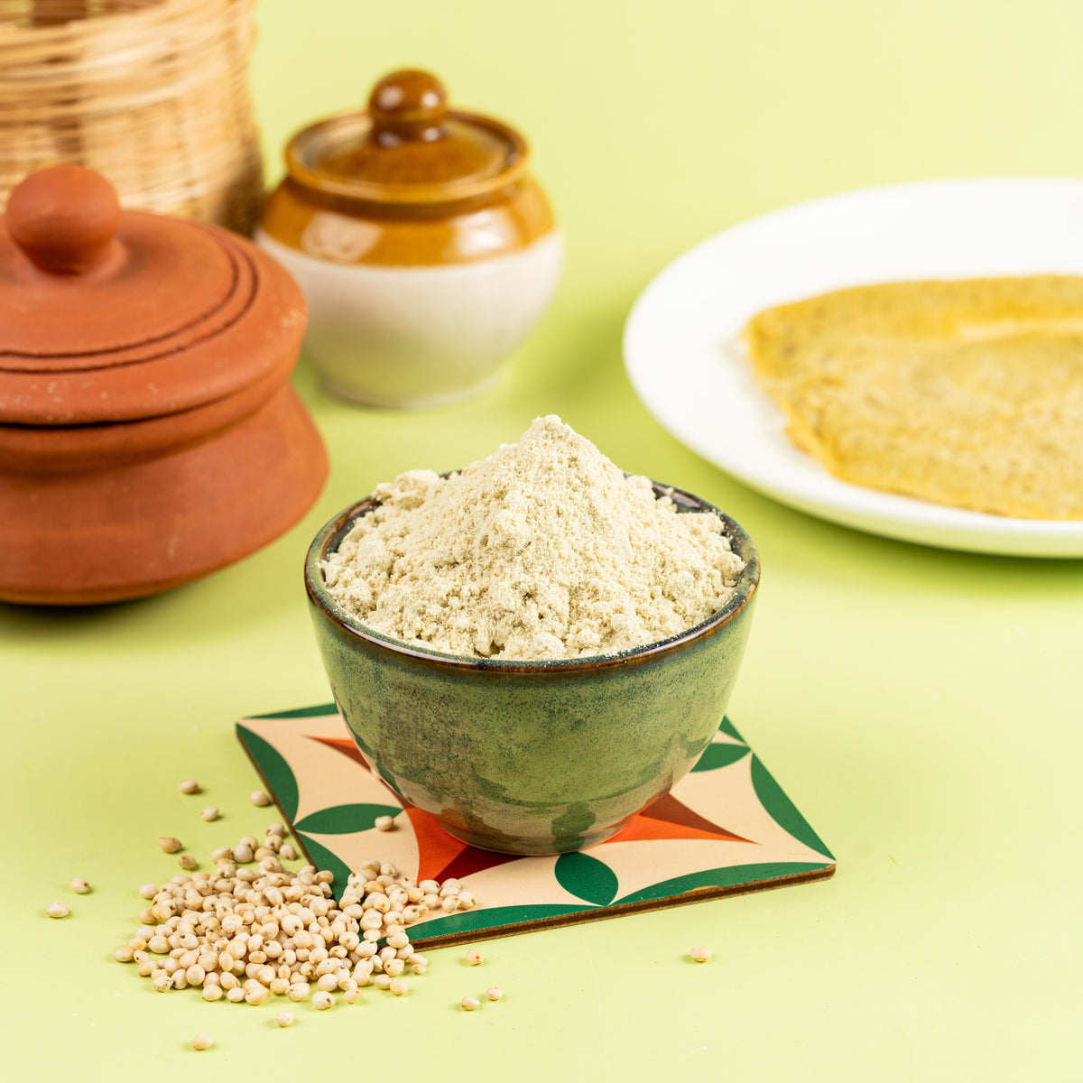 A green bowl filled with Jowar Dosa Mix, with a portion of the mix spilling out of the bowl. In the background, there is a container and a plate with Dosa prepared using the same mix.