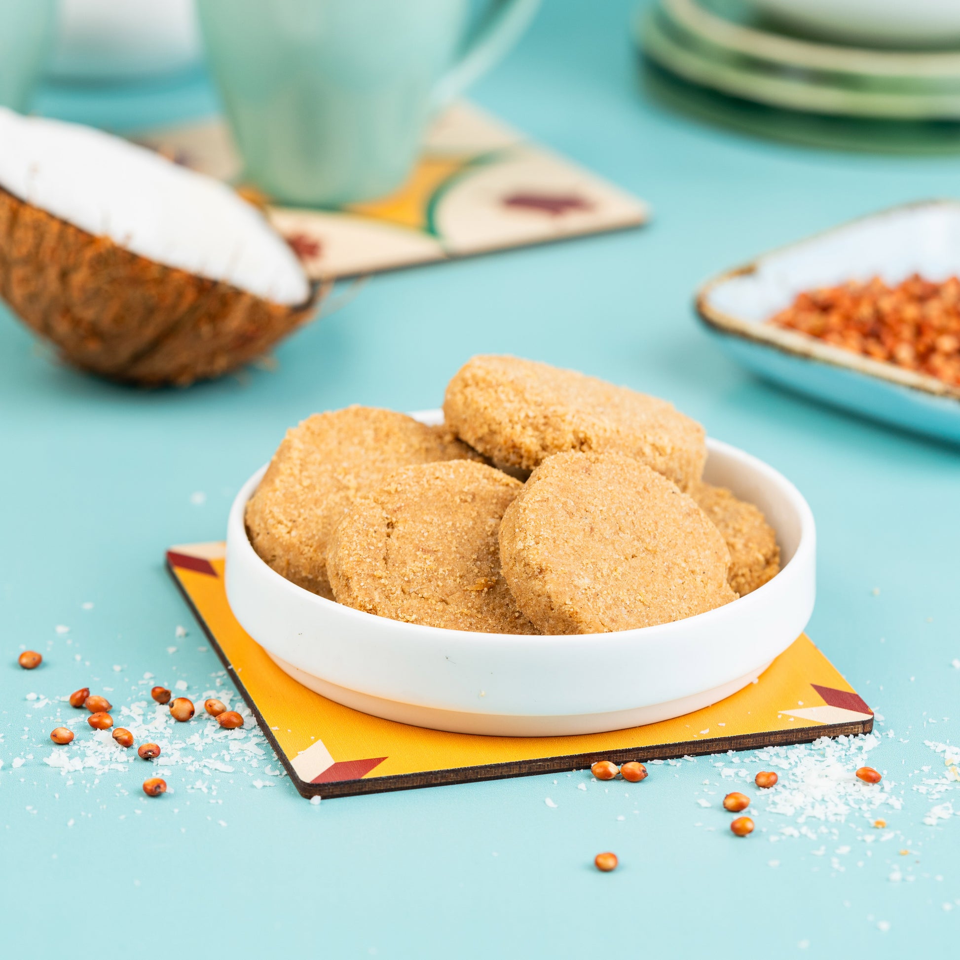 A white bowl with Jowar Coconut Cookies, presented on a colorful plate with a piece of coconut and spices in the background, all placed on a blue surface with coconut flakes scattered around.