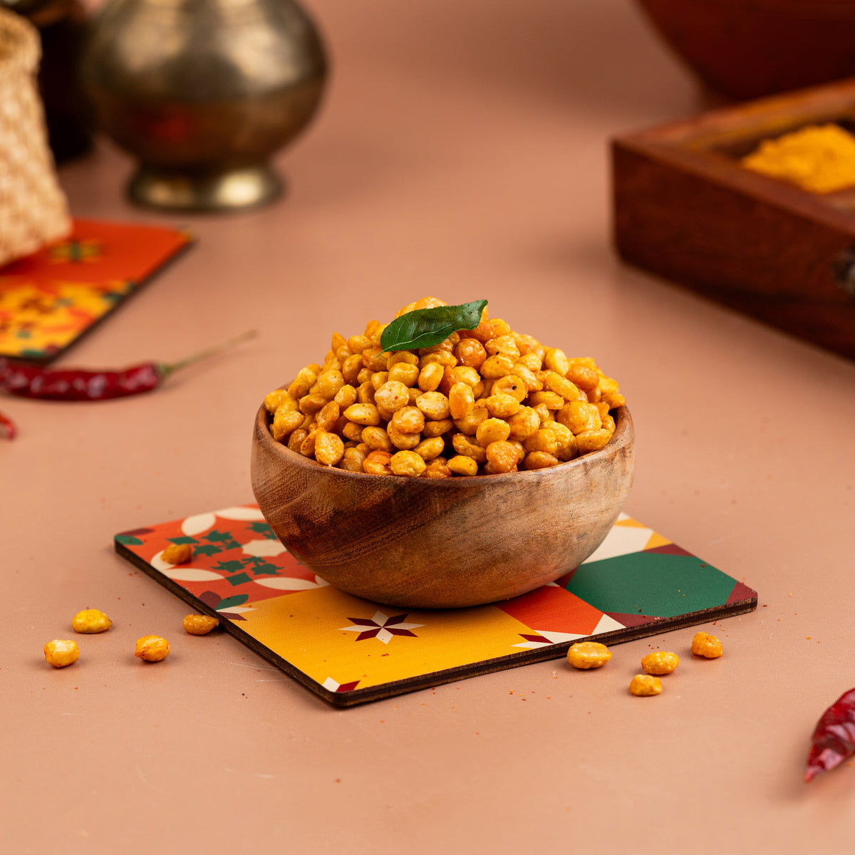 A wooden bowl filled with fried gram dal (kadalai paruppu) sprinkled with spices, placed on a colorful napkin with a few scattered beans around, against a warm-toned background.