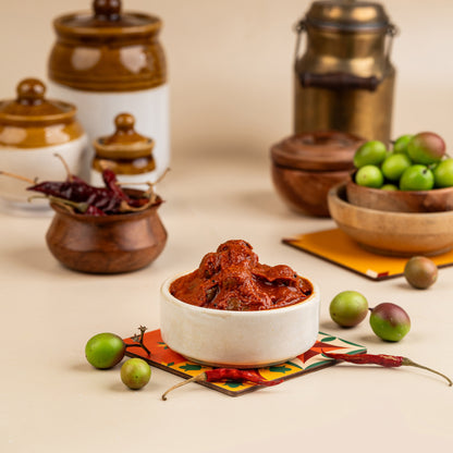 A bowl of Kalakai Pickle in the foreground with kalakai plums, spices, and traditional kitchenware in the background, all arranged on a colorful cloth.