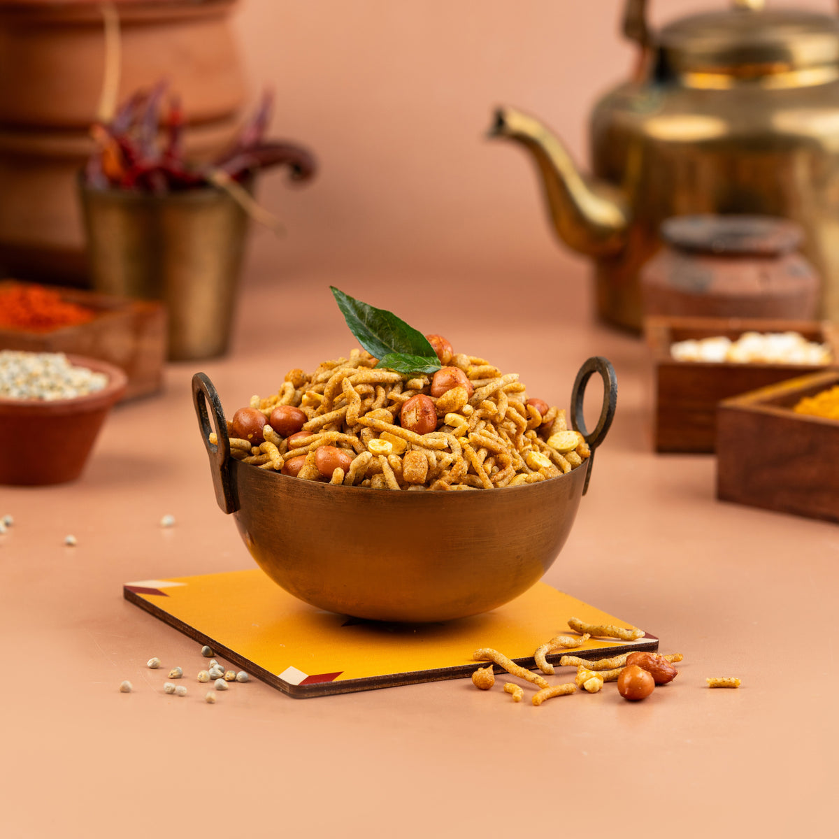 A bowl of Kambu Mixture (Pearl Millet Mixture) snack displayed on a table with decorative spices and a tea kettle in the background.