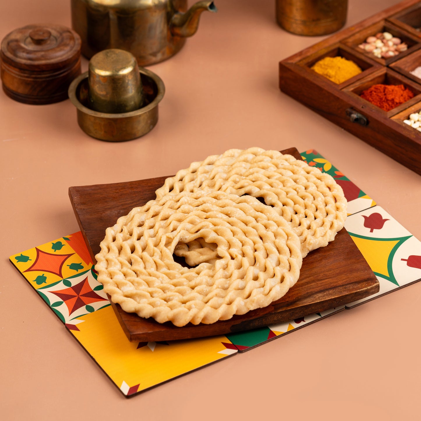 A traditional Indian snack, Murukku, presented on a colorful napkin with a wooden backdrop, alongside spices and a metal pot.