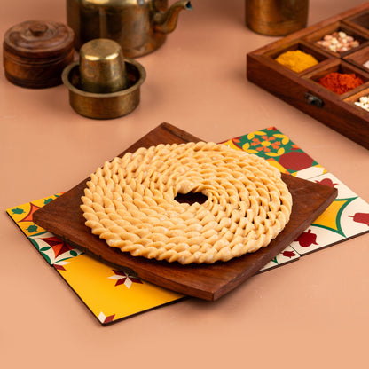 A circular, crispy murukku snack placed on a wooden board with a colorful napkin underneath, set against a backdrop featuring various kitchen utensils and spices.