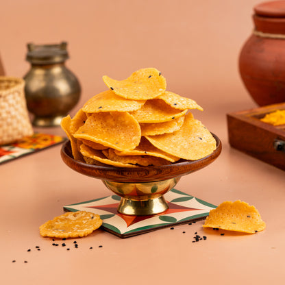 A bowl of Karaikudi Thattai (Crips) on a table with spices and a traditional Indian pot in the background.