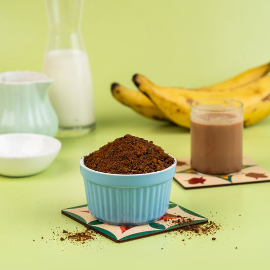 A bowl of Kerala Banana Malt powder with a prepared malt drink and bananas in the background, presented on a green surface.