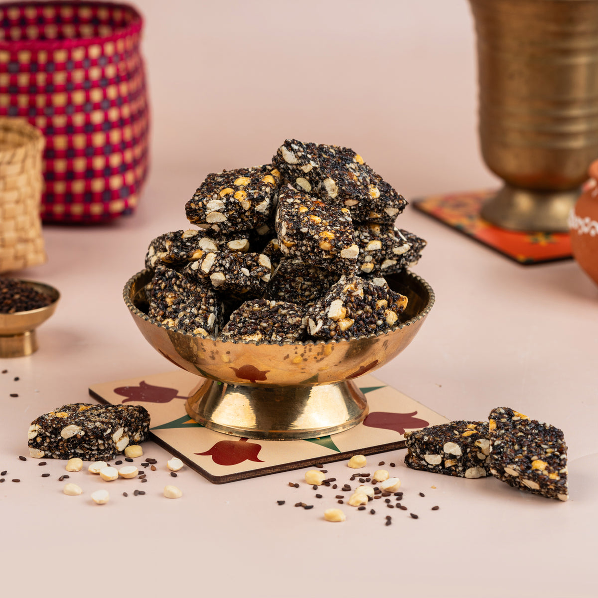 A bowl of black sesame candy on a table with some candy pieces scattered around, against a backdrop with a woven object and spices.