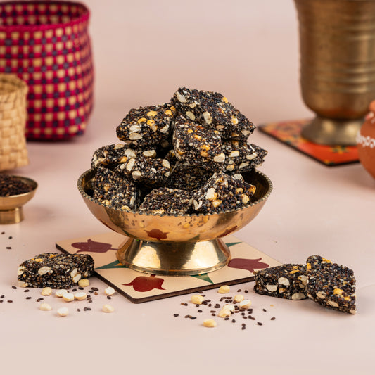 A bowl of black sesame candy on a table with some candy pieces scattered around, against a backdrop with a woven object and spices.