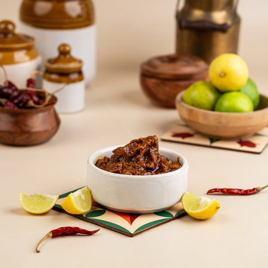 A bowl of lemon pickle in a white bowl with lemon slices and red chili peppers on the side, with a background featuring various fruits and condiments.