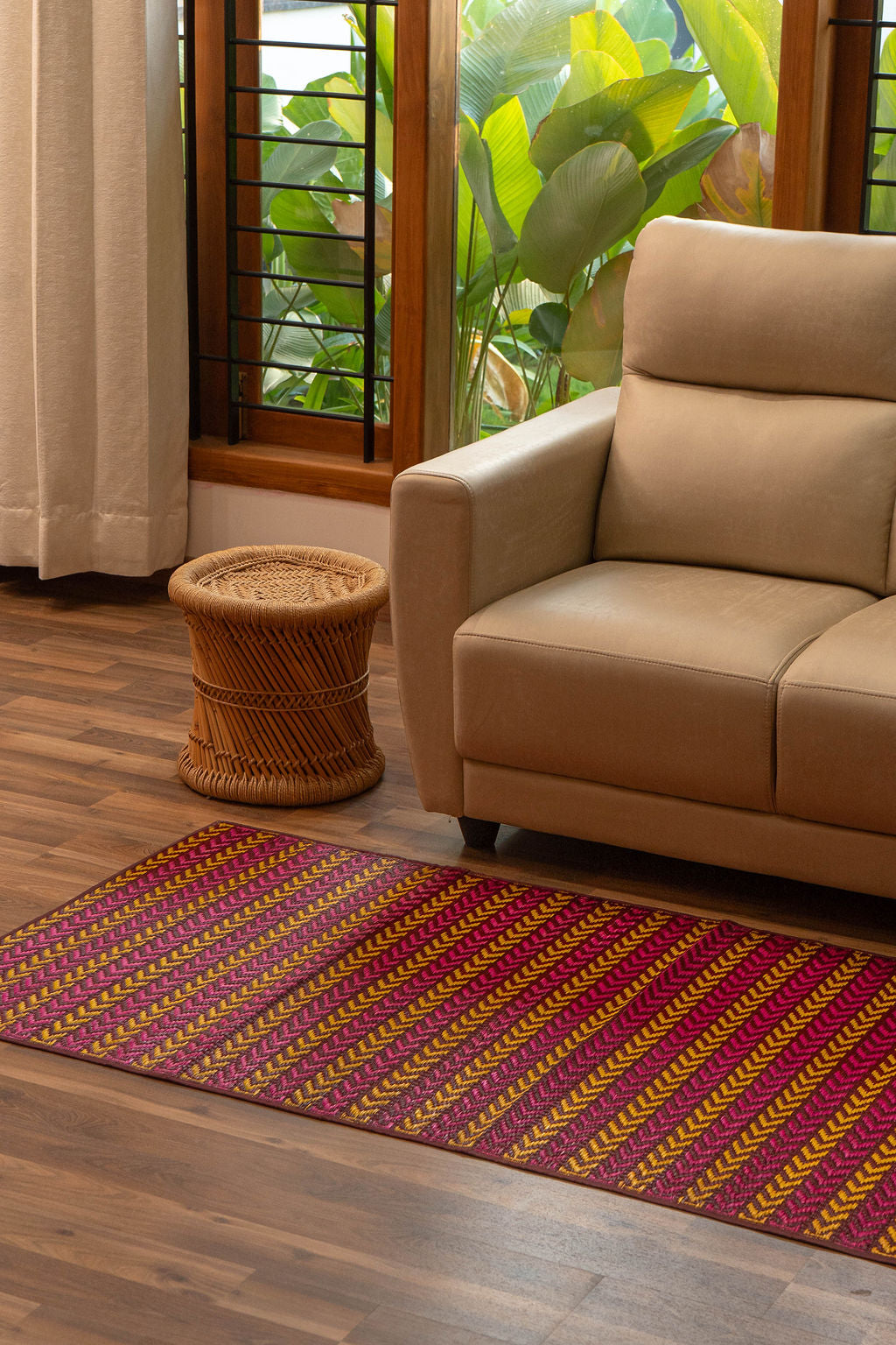 Beige sofa with a patterned Pattamadai Pai on a wooden floor, next to a window with plants.