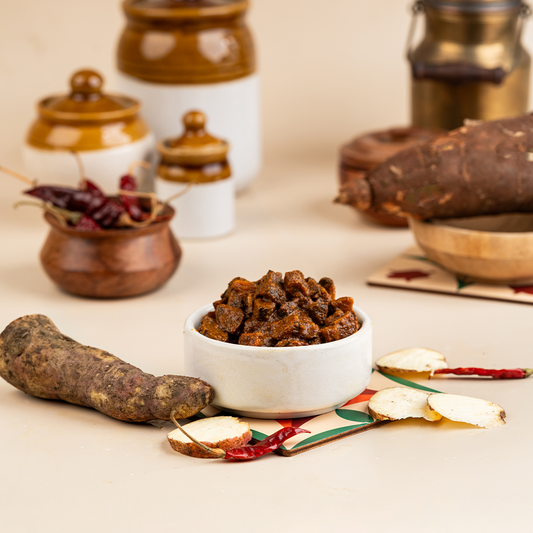 A bowl of magani kizhangu (sweet potato) pickle alongside whole and sliced sweet potatoes on a beige surface, with spices and a pickle jar in the background.